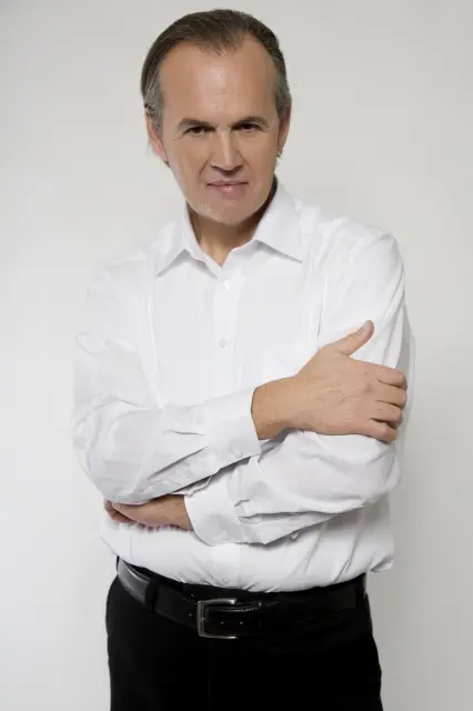 Portret Gia Suriana, u bijeloj košulji s prekrštenim rukama, ozbiljan izraz lica.